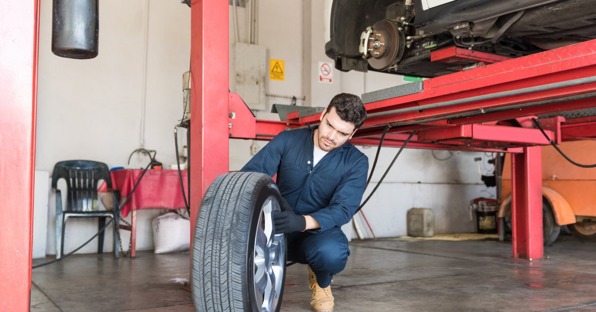 A mechanic in an auto garage is rolling a tire. Next to him is a red four-post auto lift holding up a white car.